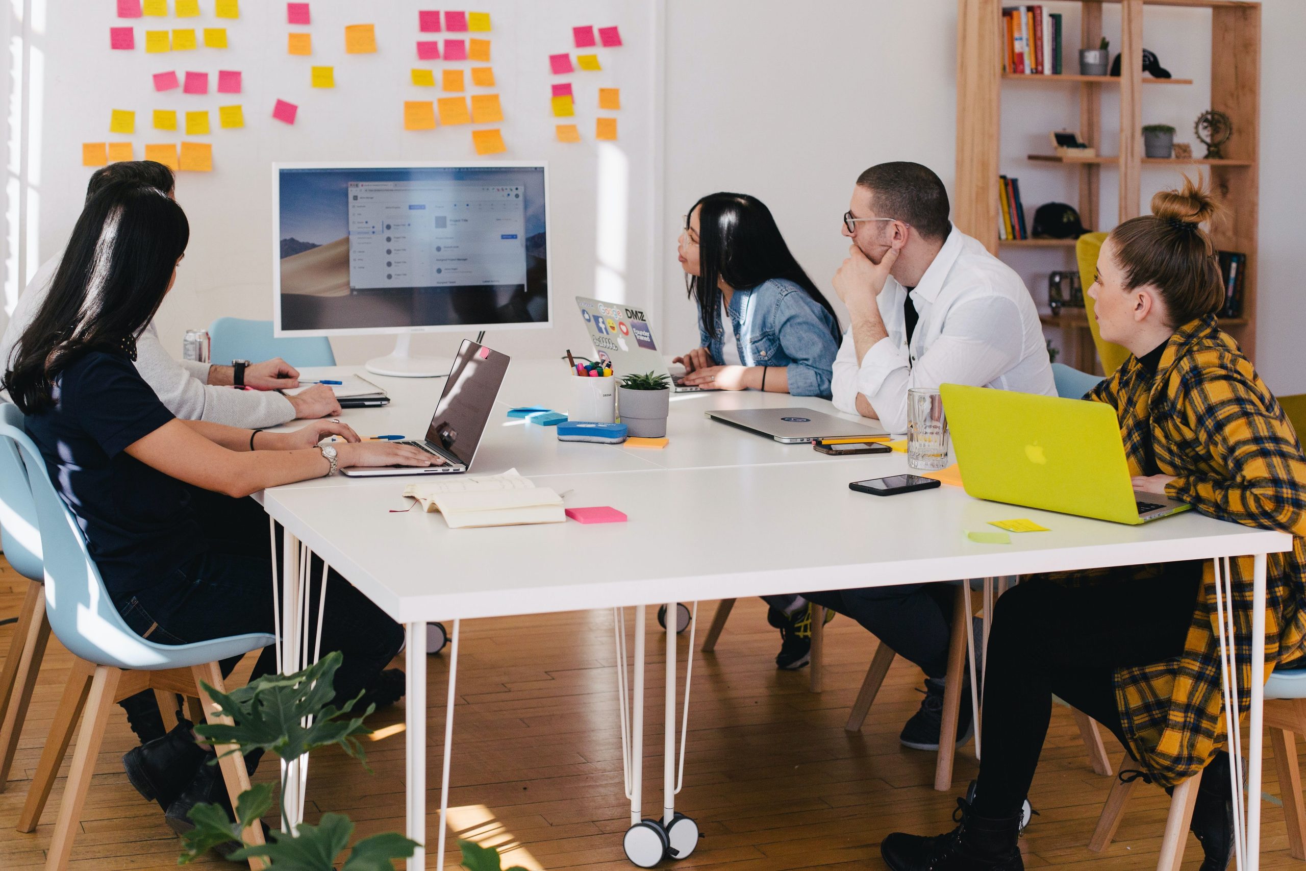 A group of people sat around a table, problem solving together. There are post-it notes on the wall and they are looking at a big, shared computer screen in front of them.